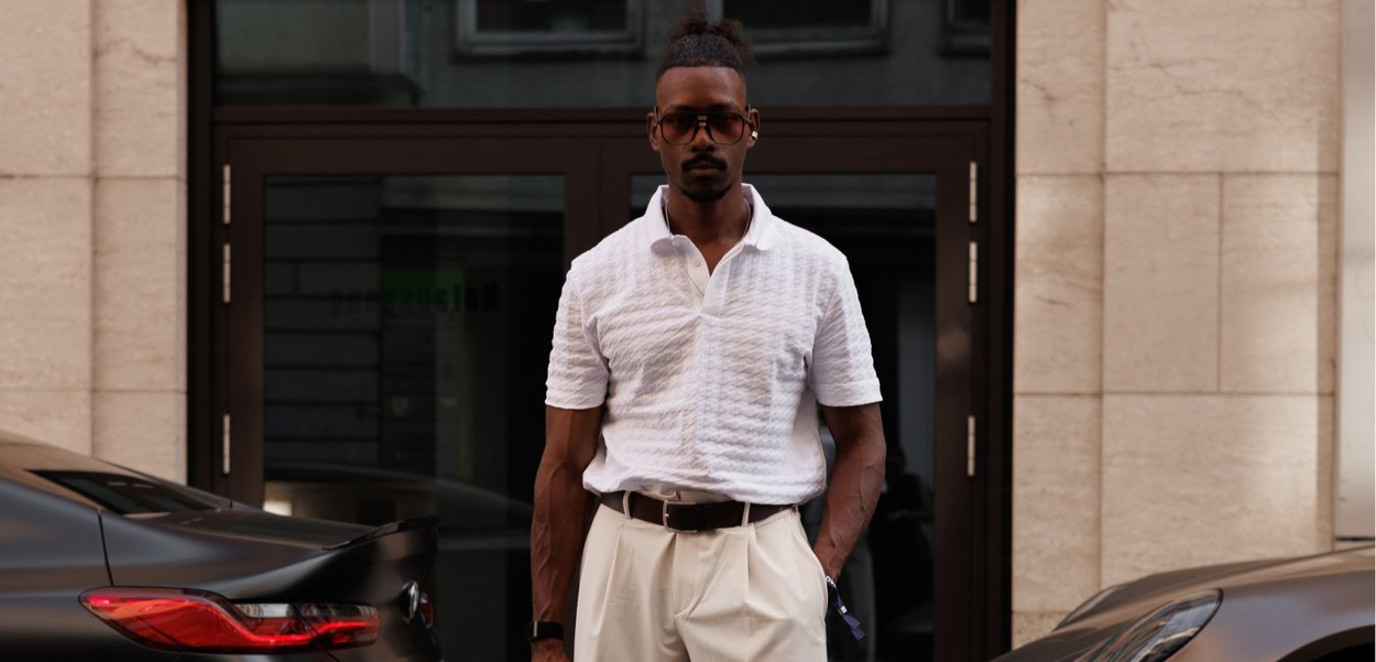 White textured polo shirt, cream trousers, dark belt, sunglasses, standing between two black cars outside a building with light-colored stone.
