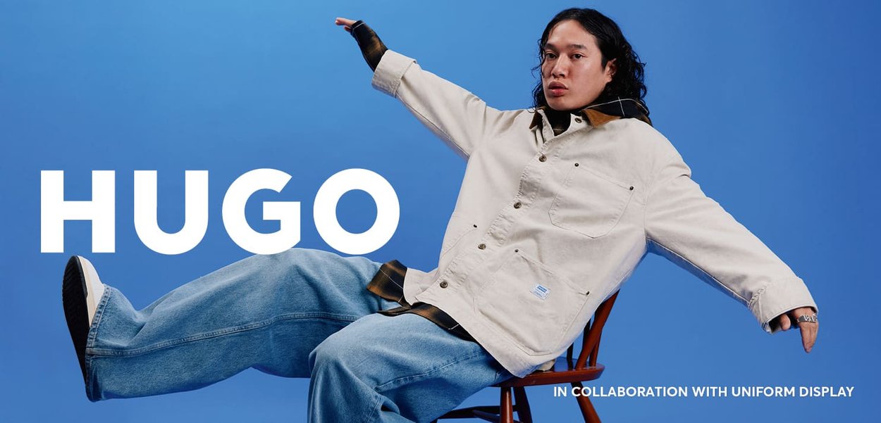 Beige jacket with a collar, featuring large buttons and pockets. Loose blue jeans and black shoes. Model poses on a chair against a blue background.