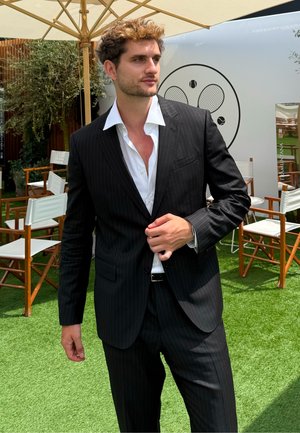 Men's black pinstripe suit featuring a structured jacket, notch lapels, and matching trousers, worn over a white shirt. Green grass background.