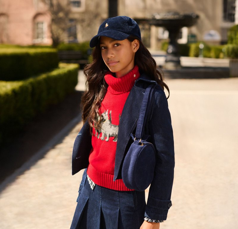 Giacca in corduroy blu, maglione a collo alto rosso con motivo animale, cappellino blu navy con logo, e borsa rotonda blu navy; abbinato a una gonna plissè.
