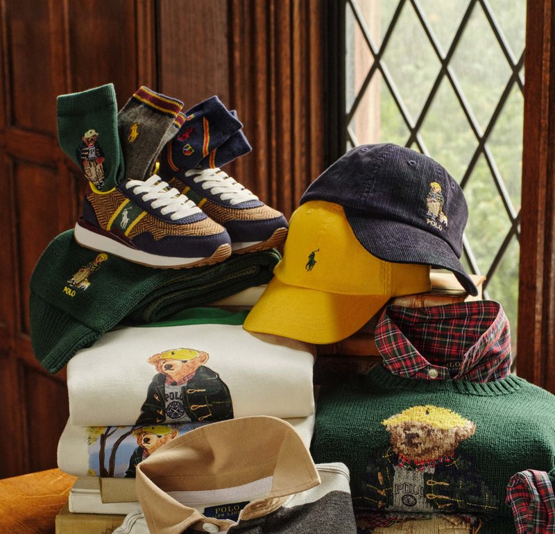 Stapel von Polo Ralph Lauren Bekleidung mit Bärenmotiven, einschließlich Pullovern, Socken, Turnschuhen mit Socken und zwei Baseballmützen in der Nähe eines Fensters.