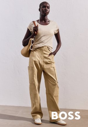 Pantaloni cargo beige con tasche laterali e una vestibilità rilassata, abbinati a una maglietta beige chiaro e scarpe da ginnastica. Gli accessori includono una borsa intrecciata e occhiali da sole.
