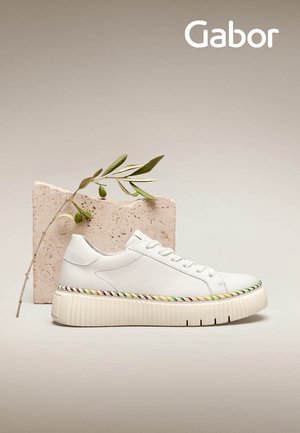 White leather sneaker with a thick, textured sole featuring rainbow-colored accents. Simple lacing and minimalistic design. Mounted on a stone block.