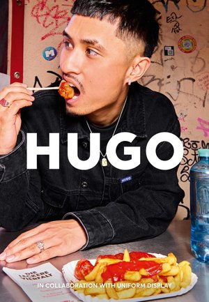 En person i en svart denimjacka med en blå etikett äter mat med pinnar. Synliga föremål inkluderar ketchup-täckta pommes frites och en dryck.