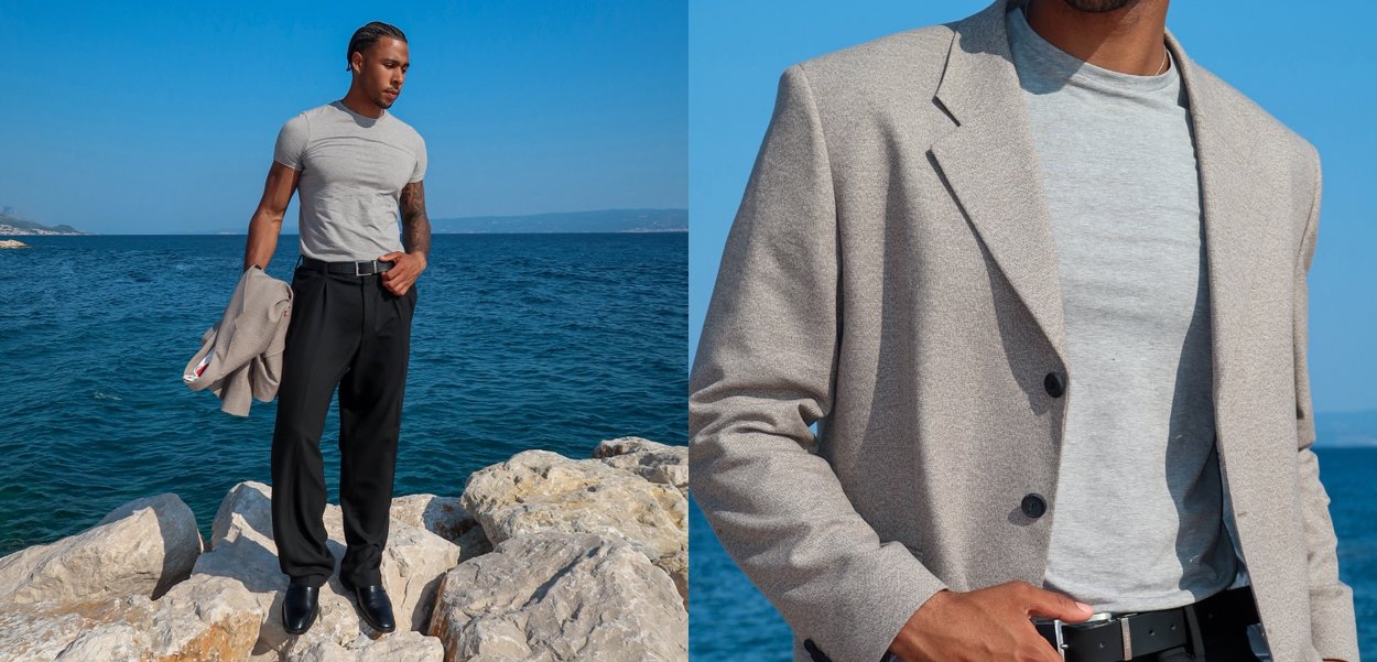 Gray fitted t-shirt, black trousers, and a light beige blazer. Blazer features two black buttons, standing on rocky shore by the blue sea.