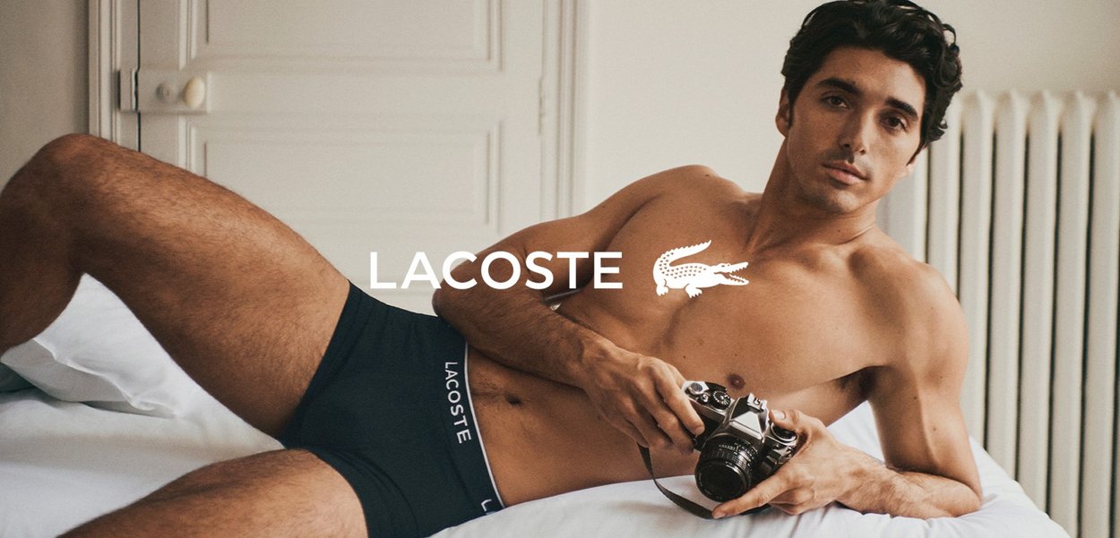 Schwarze, enganliegende Boxershorts mit einem weißen Logo, kombiniert mit einer Vintage-Kamera, vor einem hellen Hintergrund und weicher Bettwäsche.