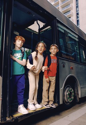 Three children with backpacks stand at the open door of a gray school bus on a city street, preparing to step off.