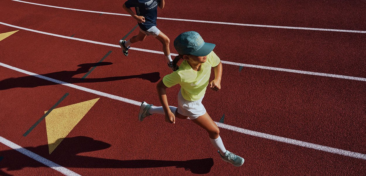 Twee kinderen rennen op een rode atletiekbaan. De één draagt een neongele shirt en een groene pet; de ander draagt een navyshirt en shorts.