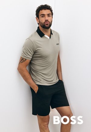 Ljust grå pikétröja med svart krage och logotyp, kombinerad med svarta shorts, står mot en vit vägg. Huden är synlig med en tatuering på armen.
