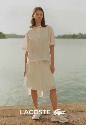 Weißes Poloshirt mit Kurzärmeln und perforiertem Design, kombiniert mit einem fließenden weißen Plisseerock. Tragen mit weißen Sneakers, vor einem Wasserhintergrund.