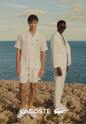 Zwei männliche Models stehen auf felsigem Gelände am Meer. Der eine trägt ein weißes, gestricktes Kurzarmshirt und Shorts; der andere trägt einen weißen Anzug und Sonnenbrille.