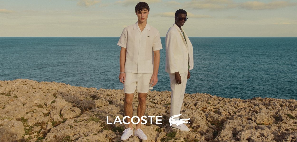 Due modelli si trovano su un terreno roccioso vicino al mare. Uno indossa una maglietta beige a maniche corte e pantaloni corti; l'altro è vestito con un completo bianco e una camicia verde.