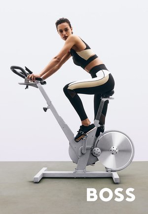 Bicicletta stazionaria con telaio argentato, grande volano e sella regolabile. Il ciclista indossa abbigliamento sportivo nero e oro. Superfici e manopole lisce.