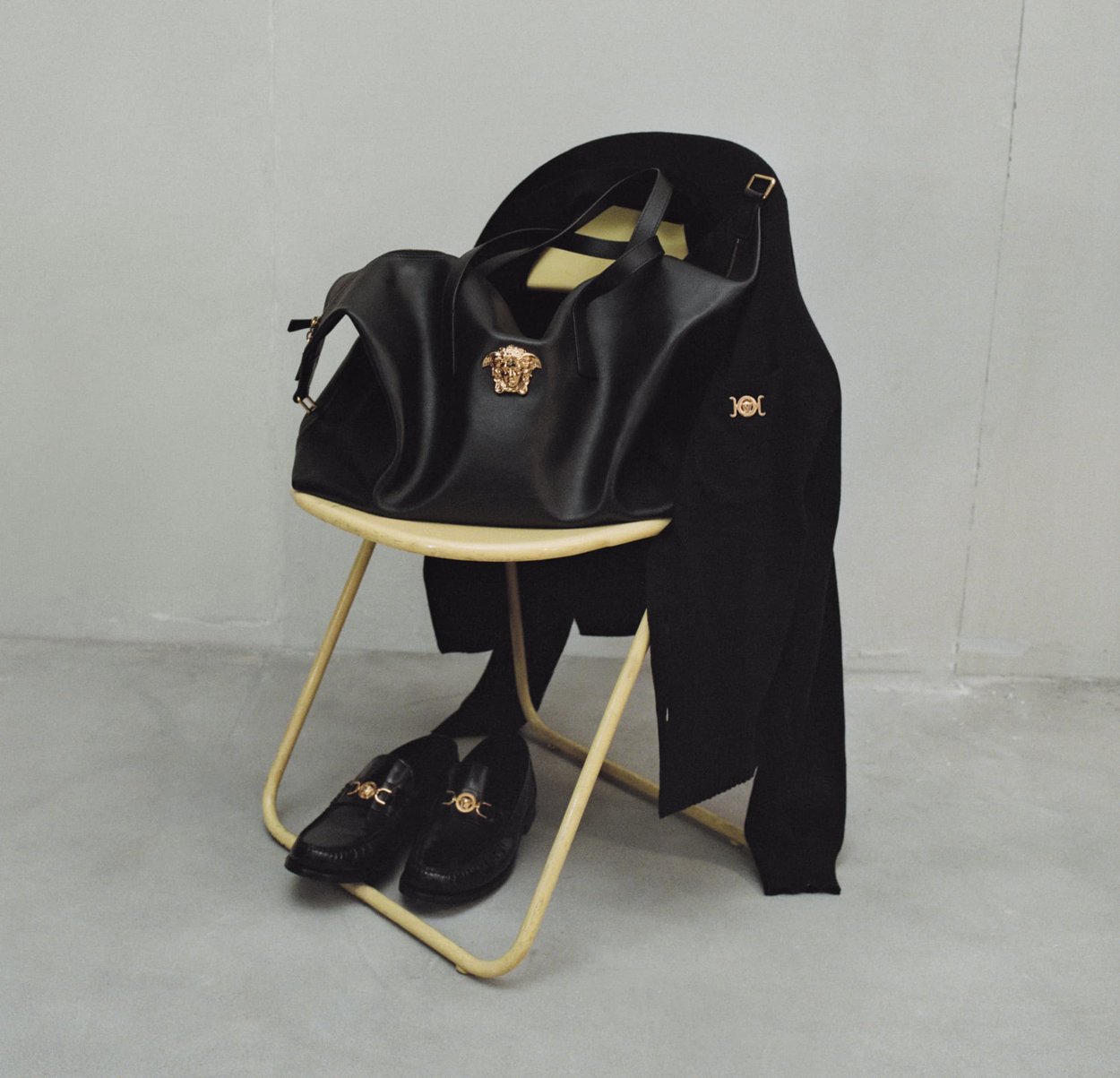 Sac fourre-tout en cuir noir avec un emblème méduse doré, posé sur une chaise teintée de jaune. Chaussures noires avec accents dorés et un manteau noir drapé à proximité.