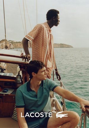 Zwei Männer in einem Boot; einer trägt ein gemustertes Korallenshirt und der andere ein einfarbiges tealfarbene Polo. Natürliches Licht hebt ihre Kleidung und die Umgebung hervor.