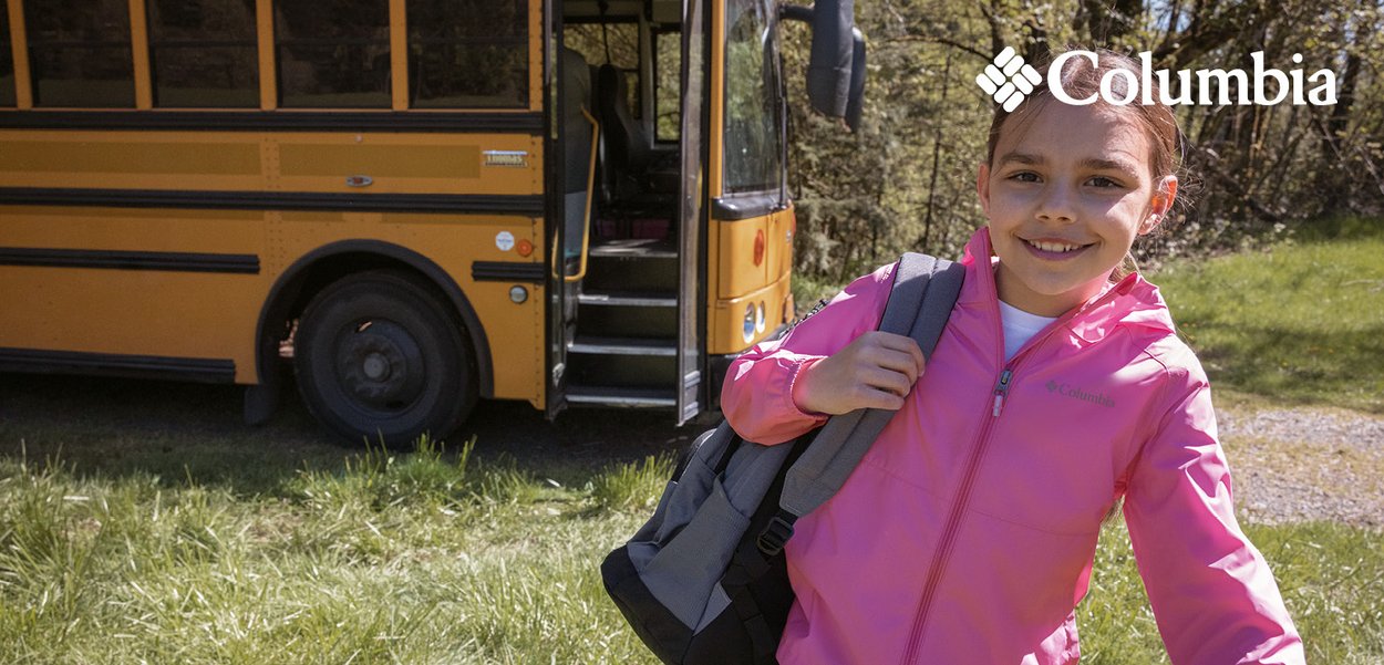 Leggera giacca rosa con zip frontale e logo, abbinata a uno zaino grigio. Sullo sfondo, un autobus giallo e erba verde.