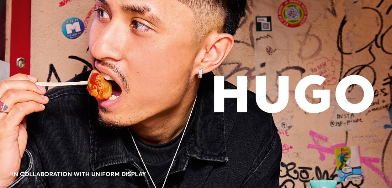 Ein Mann mit kurzen, stylischen Haaren isst mit Essstäbchen und trägt eine schwarze Jeansjacke. In fetter Schrift steht "HUGO" vor einem Graffiti-Hintergrund.