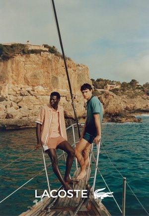 Zwei Männer stehen auf einem Holzboot. Der eine trägt ein gemustertes pinkes Hemd und Shorts, der andere ein einfarbiges grünes Hemd und blaue Shorts.