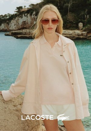 Hellrosa Polohemd, kombiniert mit einer passenden hellen Jacke, getragen über weißen Shorts. Das Modell trägt große, braungetönte Sonnenbrillen.