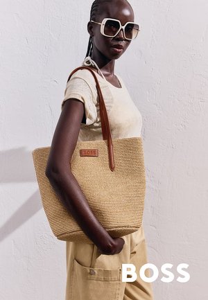 Borsa a spalla beige intrecciata con manici in pelle marrone e un piccolo logo in rilievo. Modello che indossa una camicia chiara e occhiali da sole, con uno sfondo testurizzato.