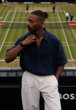 Chemise bleu marine à boutons, pantalon beige clair avec des plis, et accessoires comprenant des bagues et une montre. Fond de court de tennis.