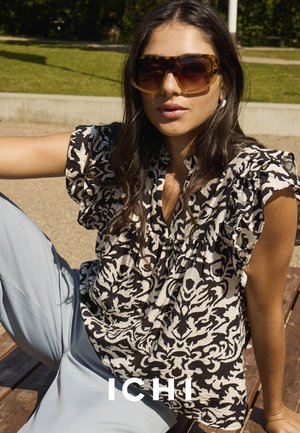 Bluse mit schwarz-weißen Mustern und Rüschemähren, kombiniert mit hellblauen Hosen, getragen von einem Modell, das auf einer Holzoberfläche sitzt.