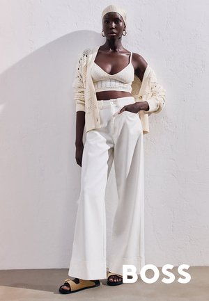 Cream-colored knitted crop top with matching cardigan, wide white trousers, and tan slide sandals. The setting features a textured wall.