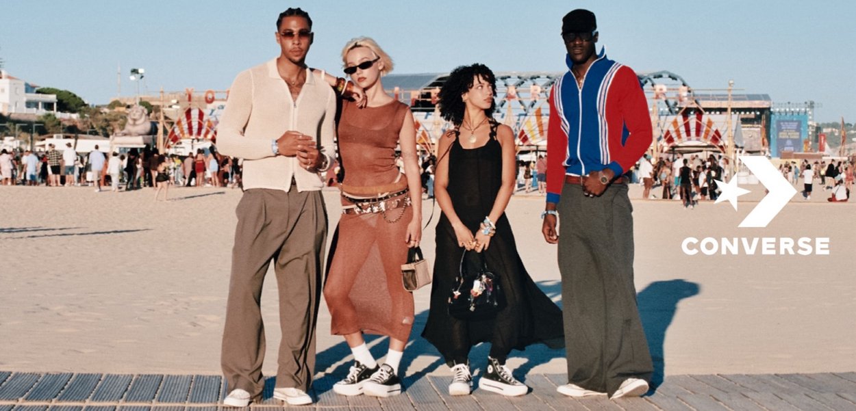 Vier individuen staan op een strand, gekleed in casual zomeroutfits. Belangrijke kenmerken zijn oversized broeken, crop tops en Converse sneakers.