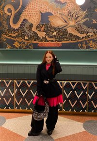 Een persoon in een zwarte blazer over een roze gelaagde jurk houdt een beaded zwarte tas vast. Op de achtergrond is een kleurrijke muurschildering van een draak te zien.