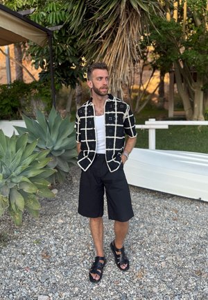 Camisa de botones de manga corta con un patrón en blanco y negro; camiseta sin mangas blanca, pantalones cortos negros, sandalias negras con tiras; fondo de vegetación al aire libre.