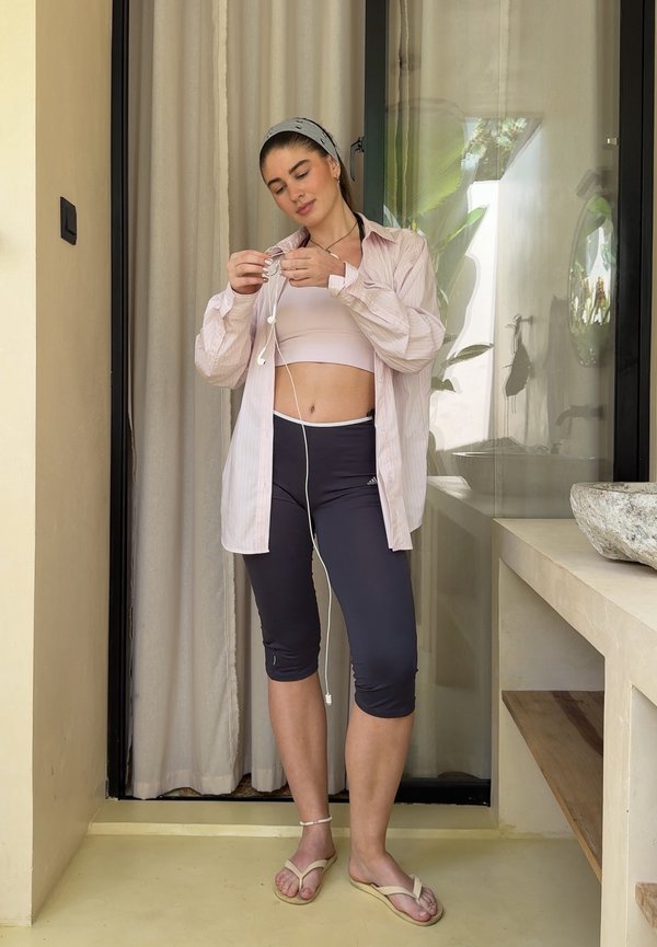 Žena v športovom oblečení a otvorenej košeli stojí vo vnútri a nastavuje si drôtové slúchadlá pri posuvných dverách s závesmi a kamenným umývadlom.