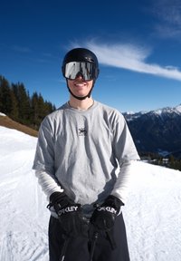 Skieur souriant portant un casque noir, des lunettes réfléchissantes, des gants Oakley gris et un shirt gris, tenant des bâtons de ski sur une montagne enneigée sous un ciel bleu.