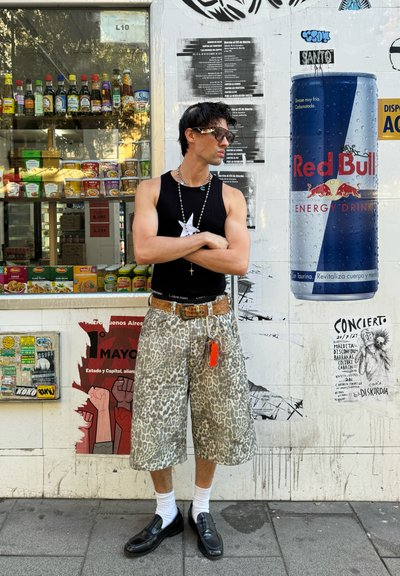 Hombre adulto con una camiseta sin mangas negra, pantalones cortos de estampado de leopardo y zapatos de piel negros con calcetines blancos. Los accesorios incluyen un collar de estrella y un cinturón.