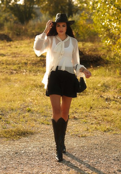 Botas de cowboy negras, blusa blanca transparente con lazo, pantalones cortos negros, bolso negro, al aire libre con vegetación de fondo.