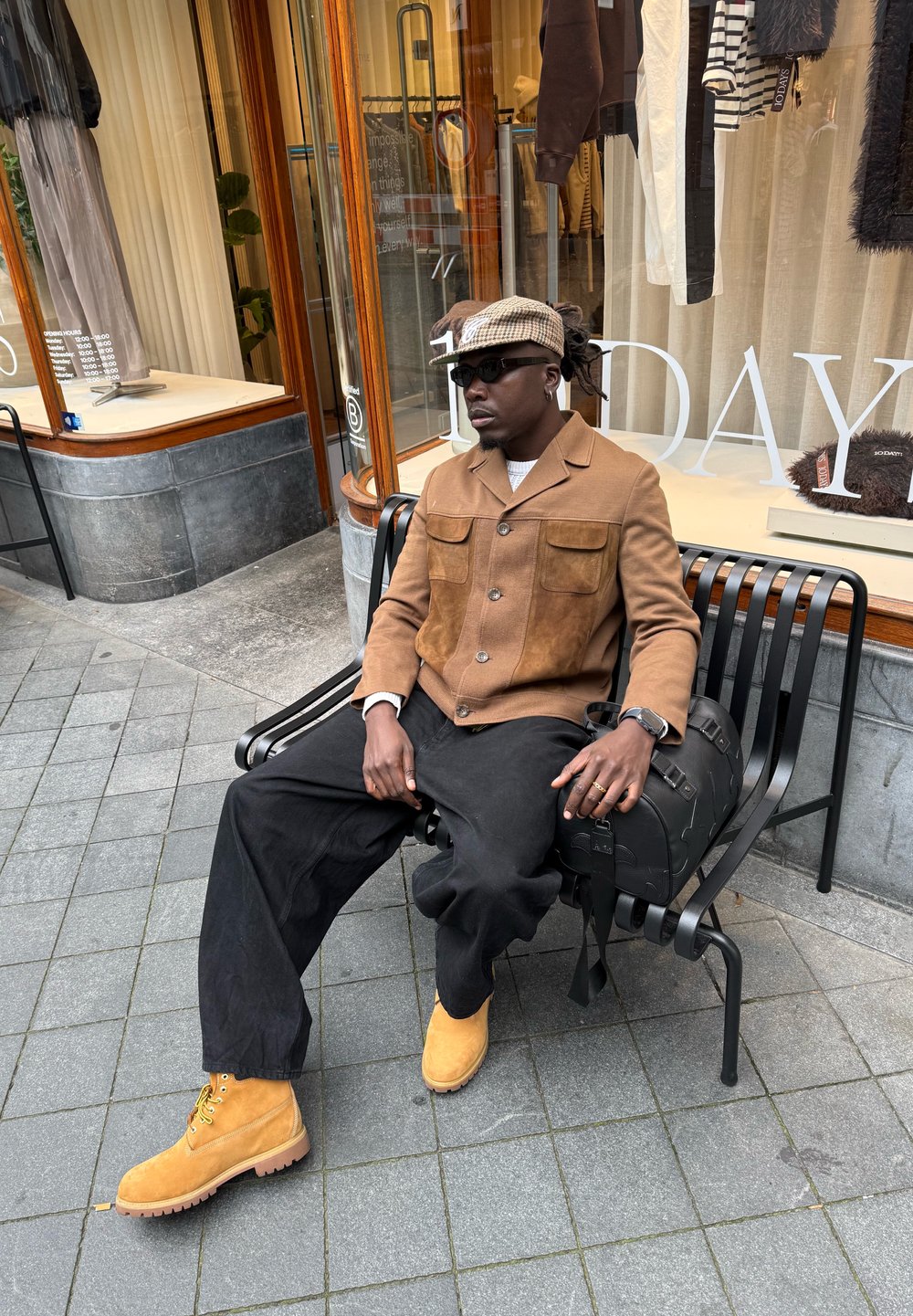Giacca a doppio tessuto marrone con due tasche, pantaloni neri a gamba larga, stivali gialli in suede e un cappello a motivo appoggiato su una panchina nera.