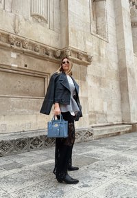 Črna usnjena jakna, siva plastena pulover, bela srajca, črne čipkaste hlače, modra torbica, črne gležnjarje, sončna očala; stoji blizu kamnite stene.