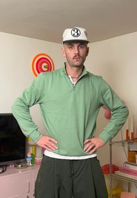 Pulover verde deschis cu fermoar până la jumătate, realizat dintr-un material moale, asortat cu pantaloni largi de culoare verde închis. Accesoriile includ o șapcă albă cu numărul negru "3".