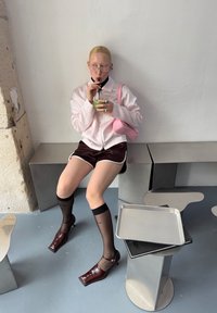 Chaqueta rosa con cremallera, pantalones cortos marrones con ribete blanco, medias negras hasta la rodilla y zapatos de tacón burdeos. Sosteniendo una bebida verde en un vaso transparente.