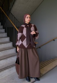 Pulover în carouri maro și bej, pantaloni maro cu picior larg, geantă asortată și ghete negre până la gleznă, plasate în fața unei scări cu balustradă de lemn.