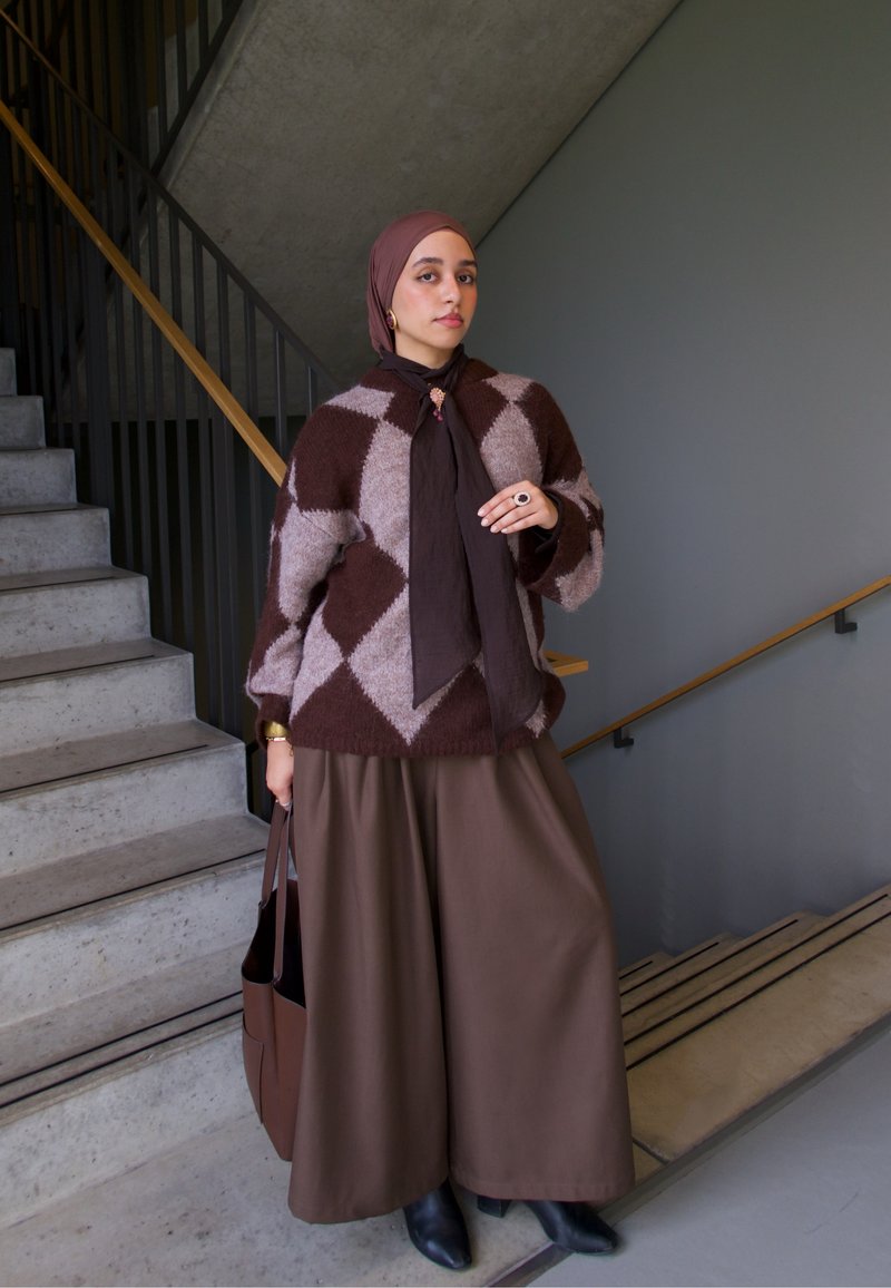 Suéter de losangos castanho e bege, calças castanhas de perna larga, bolsa combinando e botins pretos, com uma escadaria com corrimão de madeira ao fundo.