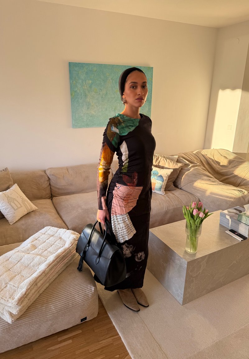 Femme en longue robe colorée et foulard, tenant un sac à main noir, debout dans un salon éclairé par le soleil avec un canapé beige et des tulipes sur une table en marbre.