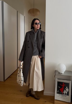 Mujer con abrigo gris oscuro, suéter gris, falda crema, zapatos marrones y gafas de sol, sosteniendo un bolso blanco con lunares dentro de una habitación con suelo de madera.