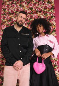 Camisa preta de botões com remendos, calças rosas, camisa listrada rosa, corpete preto, saia preta plissada e uma bolsa rosa em forma de coração.
