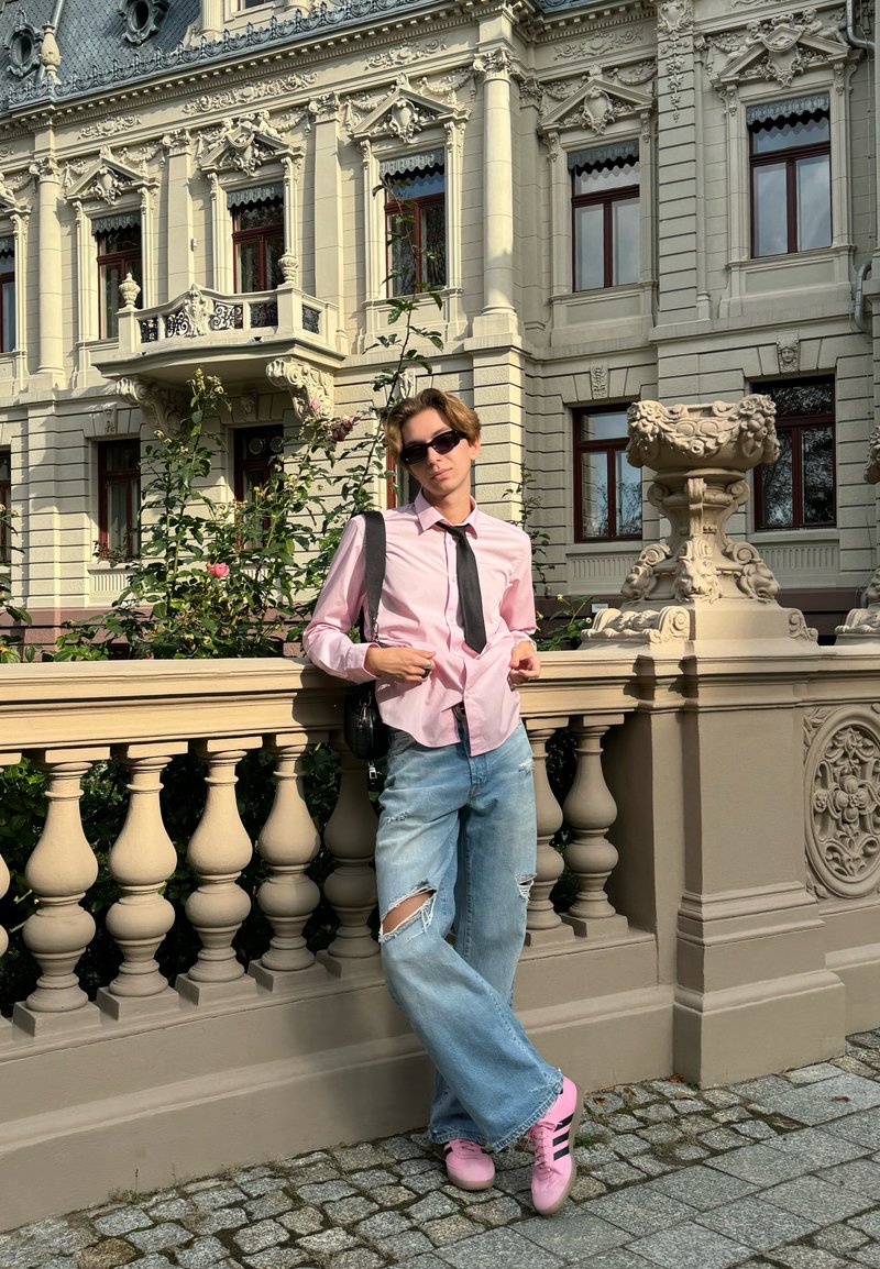 Човек, опрян о камерен парапет, носещ светло розова риза, черно вратовръзка, износени светлосини дънки и розови маратонки с черни ивици.