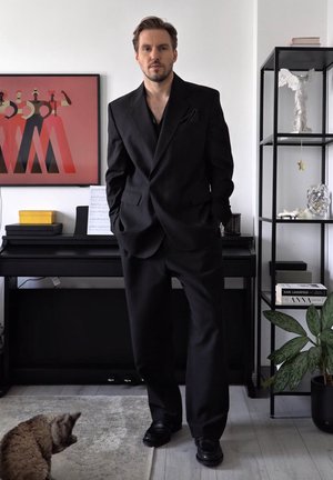 Schwarzer Anzug mit strukturierter Blazerjacke und lockeren Hosen, kombiniert mit schwarzen Schuhen. Der Hintergrund enthält abstrakte Kunst und ein schwarzes Regal mit dekorativen Gegenständen.