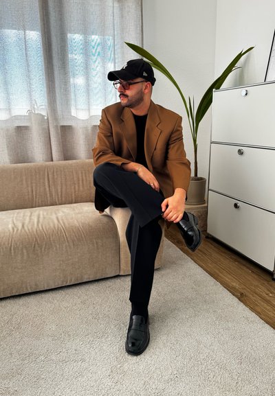 Blazer marrón de gran tamaño sobre una camisa negra, pantalones negros y zapatos negros, sentado en un sofá beige con una planta verde y un mueble cerca.