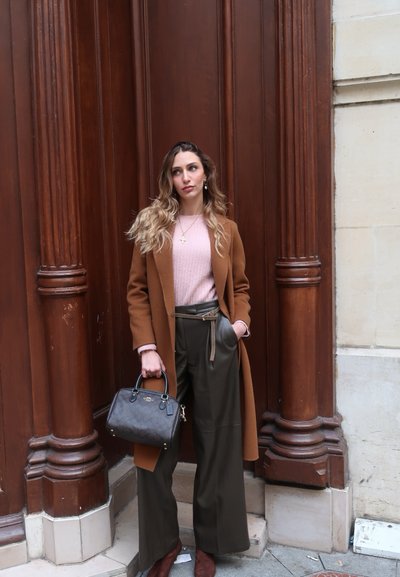Mujer con cabello largo junto a grandes columnas de madera marrón, lleva un abrigo marrón, suéter rosa, pantalones anchos oscuros y sostiene un bolso negro.