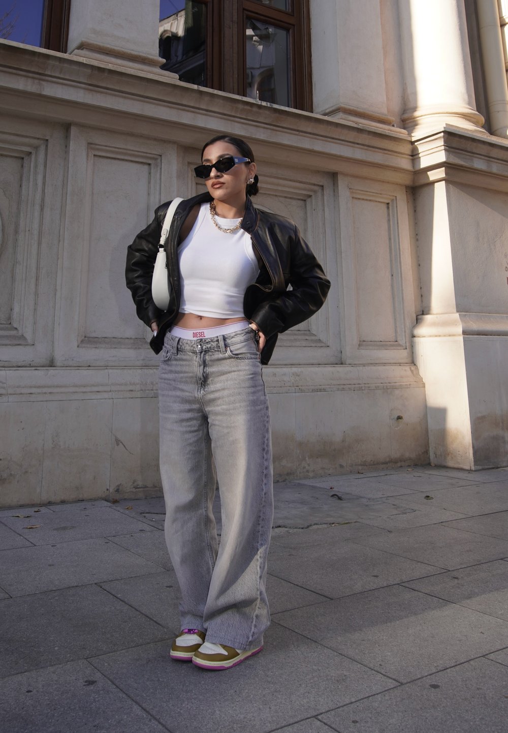 Sort læderjakke, hvid crop top, grå brede bukser. Iført solbriller, med kraftige sneakers der har grønne og pink accenter.