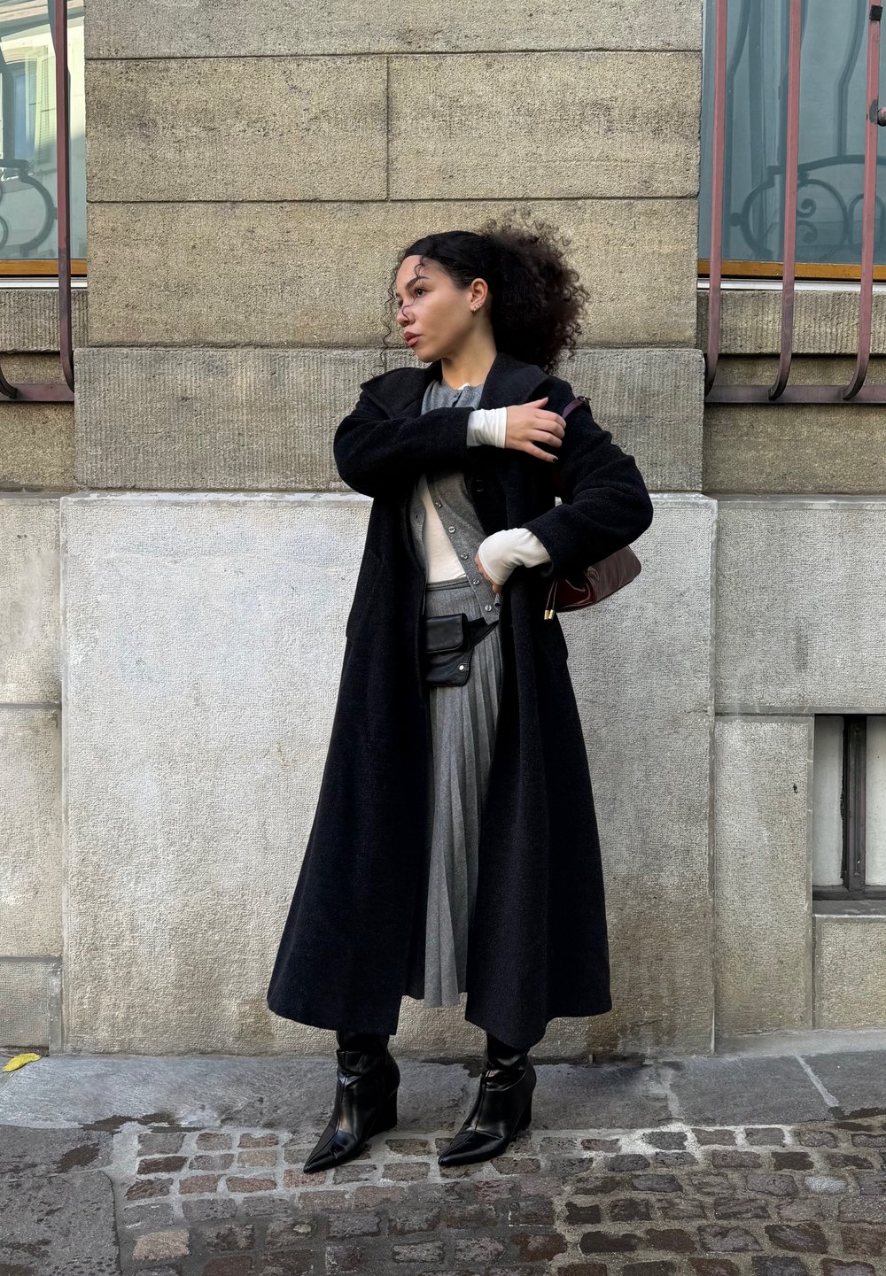 Frau mit lockigem Haar, die einen langen schwarzen Mantel, einen grauen plissierten Rock, schwarze Stiefel trägt und eine braune Schultertasche hält, während sie auf Kopfsteinpflaster steht.