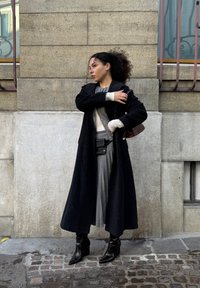 Frau mit lockigem Haar, die einen langen schwarzen Mantel, einen grauen plissierten Rock, schwarze Stiefel trägt und eine braune Schultertasche hält, während sie auf Kopfsteinpflaster steht.